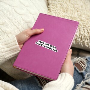 K&ouml;nnte beinhalten: Ein rosafarbenes, ledergebundenes Tagebuch mit einem wei&szlig;en Aufkleber, auf dem steht: "Schau mich nicht an, ich habe nicht f&uuml;r ihn gestimmt."