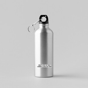K&ouml;nnte beinhalten: Eine silberne Wasserflasche mit schwarzem Deckel und einer Trageschlaufe. Die Flasche hat einen Aufkleber mit dem Text "Schau mich nicht an, ich habe nicht f&uuml;r ihn gestimmt".
