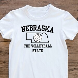 May include: White t-shirt with black text that reads "Nebraska The Volleyball State" and an outline of the state of Nebraska with a volleyball inside.