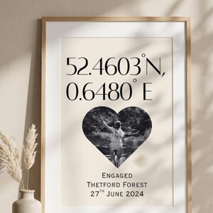 May include: A framed wall print featuring the coordinates 52.4603 N, 0.6480 E in black text. Below the coordinates is a heart-shaped black and white photograph of a person on a bicycle. The text ENGAGED THETFORD FOREST 27TH JUNE 2024 appears below.