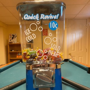 May include: A blue and silver gumball machine with a clear glass top. The machine has a sign that says "Quick Revive! 10¢" and "ICE COLD" and "SOLD HERE". The machine is filled with colorful gumballs.