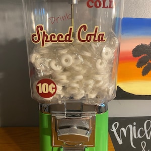 May include: A vintage green gumball machine with a clear plastic container filled with white gumballs. The machine has a chrome coin slot and a chrome lever. The machine has a sign that says "Drink Speed Cola" and "Ice Cold" in red lettering. The machine also has a sign that says "10c".