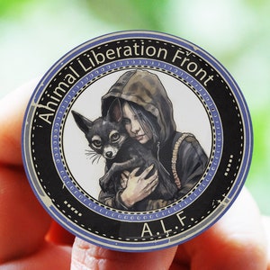 Op de afbeelding: Een ronde knop met een zwart-witte afbeelding van een persoon in een hoodie die een kleine zwart-witte hond vasthoudt. De tekst "Animal Liberation Front" staat in een cirkel rond de afbeelding, met "A.L.F." eronder.