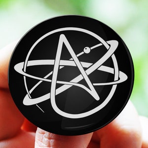 Op de afbeelding: Een zwarte knop met een wit gestileerd atoomsymbool. Het atoomsymbool is een cirkel met een ster erin en een ring eromheen.