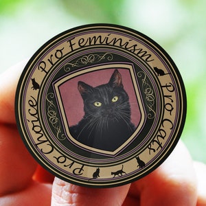 Puede incluir: Un botón redondo de color dorado con texto negro que dice "Pro Choice Pro Cat Pro Feminism". El botón presenta un gato negro con ojos verdes en un escudo rosa.