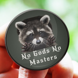 Puede incluir: Un botón redondo con fondo negro y una cara de mapache en el centro. El mapache está mirando a la cámara con las patas levantadas. El texto "No Gods No Masters" está escrito en blanco debajo del mapache.