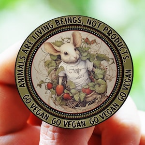 Op de afbeelding: Een ronde, gouden pin met een wit konijn dat een shirt draagt met de tekst "Go Vegan" in een mand met rode aardbeien. De pin heeft de tekst "Animals are living beings, not products. Go Vegan Go Vegan Go Vegan Go Vegan" rond de rand.