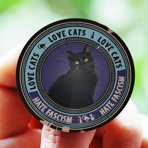 May include: A black and white round sticker with a black cat in the centre. The sticker has the text "Love Cats Love Cats Hate Fascism Hate Fascism" around the edge.