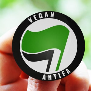 May include: A black and white button with a green flag and the text "Vegan Antifa" in white.
