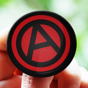 Pode incluir: Um botão redondo preto com um símbolo "A" vermelho dentro de um círculo vermelho, circundado por um anel preto. O botão é segurado contra um fundo verde desfocado.