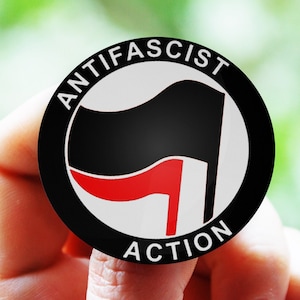 Può includere: Un bottone bianco e nero con il testo "ANTIFASCIST ACTION" in un cerchio attorno a una bandiera nera con una striscia rossa.