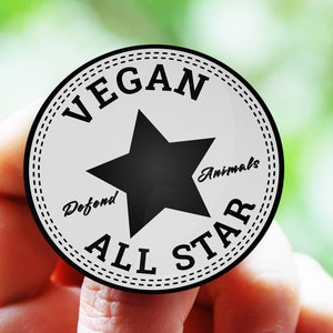 Può includere: Un adesivo rotondo bianco e nero con il testo "Vegan All Star" e una stella nera al centro. Il testo "Defend Animals" è scritto intorno alla stella.