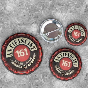 May include: Three round pins with the text "Anti-Fascist 161 Gegen Nazis" in black and white on a red background. The pins are on a grey textured surface.
