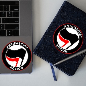 May include: A black and white sticker with a red symbol and the text "Anti-Fascist Action" on a silver laptop and a black notebook.