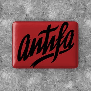 Może przedstawiać: Czerwony kwadratowy przycisk z napisem "antifa" napisanym czarnym, ozdobnym pismem.