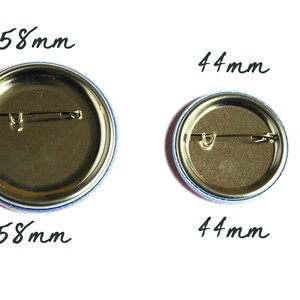 May include: Two round metal button pins with gold backs and safety pins. The larger pin is 58 mm in diameter and the smaller pin is 44 mm in diameter.