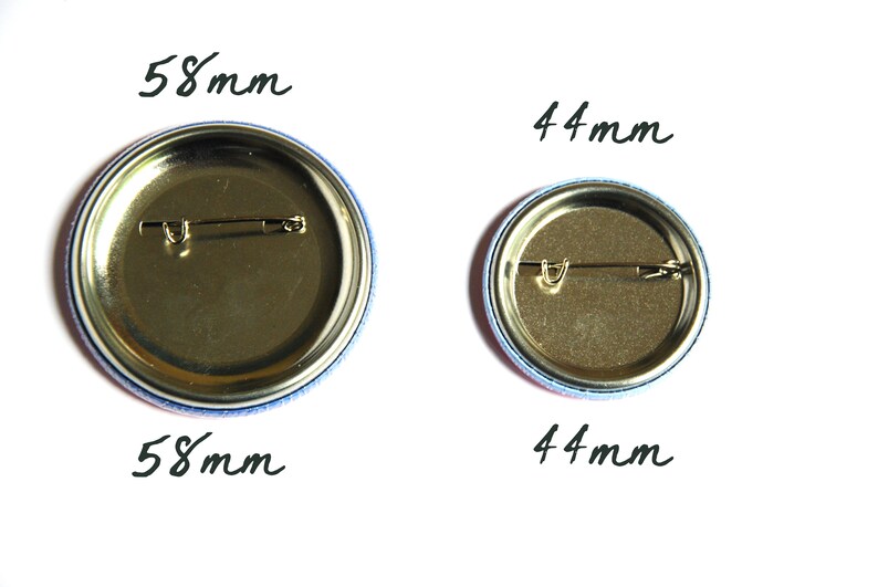 May include: Two round metal button pins with gold backs and safety pins. The larger pin is 58 mm in diameter and the smaller pin is 44 mm in diameter.