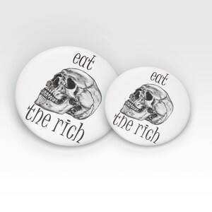 May include: Two white round buttons with black text and a black and white skull graphic. The larger button says "eat the rich" and the smaller button says "eat the rich".