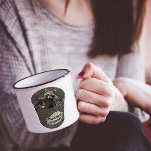 May include: White ceramic mug with a black rim and handle. The mug features a cartoon raccoon sitting in a bin with the text "It's called a trash can, not trash candy".
