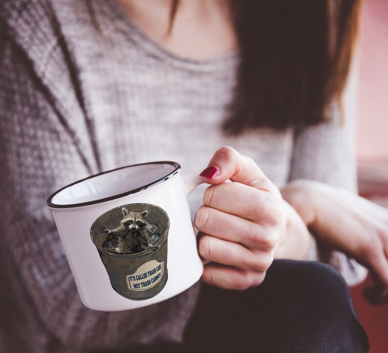 May include: White ceramic mug with a black rim and handle. The mug features a cartoon raccoon sitting in a bin with the text "It's called a trash can, not trash candy".
