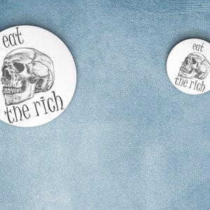 May include: Two white round buttons with a black and white skull graphic and the text "eat the rich". The larger button is on the left and the smaller button is on the right.