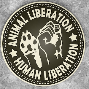 Puede incluir: Una pegatina circular en blanco y negro con el texto "Animal Liberation Human Liberation" alrededor del borde. Un puño en alto y una huella de pata están en el centro de la pegatina.