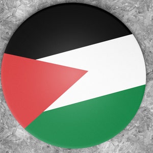 Può includere: Un bottone rotondo con la bandiera palestinese. La bandiera è nera, bianca, verde e rossa. I colori sono disposti in strisce orizzontali con un triangolo rosso nell'angolo in alto a sinistra.