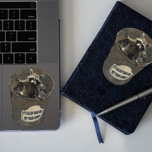 May include: A sticker of a raccoon in a trash can with the text "It's called trash can, not trash cannot." The sticker is on a silver laptop. A blue notebook with a floral pattern and a silver pen is next to the laptop.