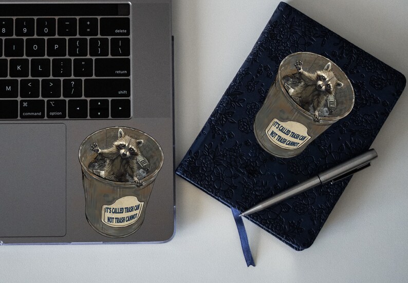 May include: A sticker of a raccoon in a trash can with the text "It's called trash can, not trash cannot." The sticker is on a silver laptop. A blue notebook with a floral pattern and a silver pen is next to the laptop.