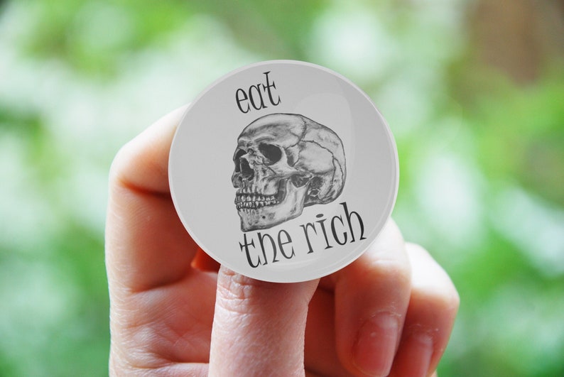 May include: A white button with a black and white skull and the text "eat the rich" printed on it.
