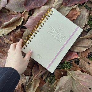 Peut inclure: Un agenda à spirales de couleur crème avec des spirales dorées et le texte "THE PLANNER DES CM" en relief sur la couverture. Une bande élastique rose maintient l'agenda fermé. L'année "2020" est imprimée.