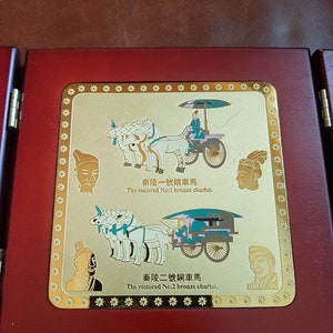 May include: A wooden plaque with a gold-colored frame and two images of bronze chariots. The text on the plaque reads "秦陵一號銅車馬 The restored No:1 bronze chariot." and "秦陵二號銅車馬 The restored No:2 bronze chariot."
