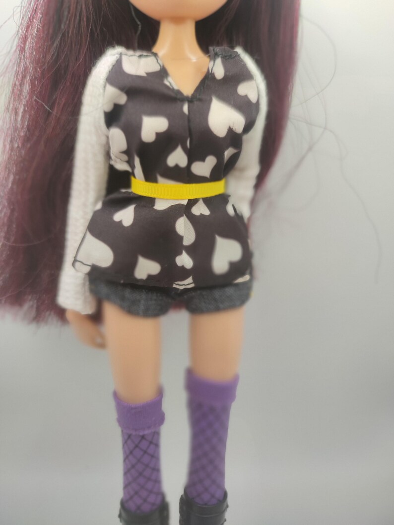 Pode incluir: Uma boneca vestindo um top preto e branco com estampa de cora&ccedil;&atilde;o com cinto amarelo, mangas brancas e shorts cinza. A boneca tem cabelo roxo, meias de rede roxas e sapatos pretos. &Eacute; uma boneca fashion.