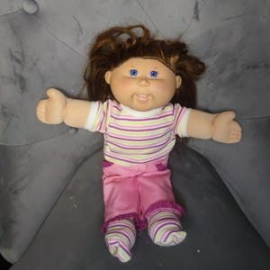 Peut inclure: Une poupée Cabbage Patch Kids avec des cheveux bruns et des yeux bleus. La poupée porte une chemise rayée rose, verte et blanche, un pantalon rose et des chaussettes rayées. La poupée est sur une surface grise.