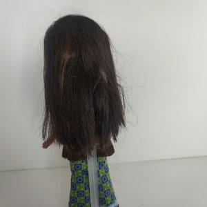 May include: A brown-haired doll wearing a brown vest, green and blue checkered pants with pink and blue trim, and pink shoes.