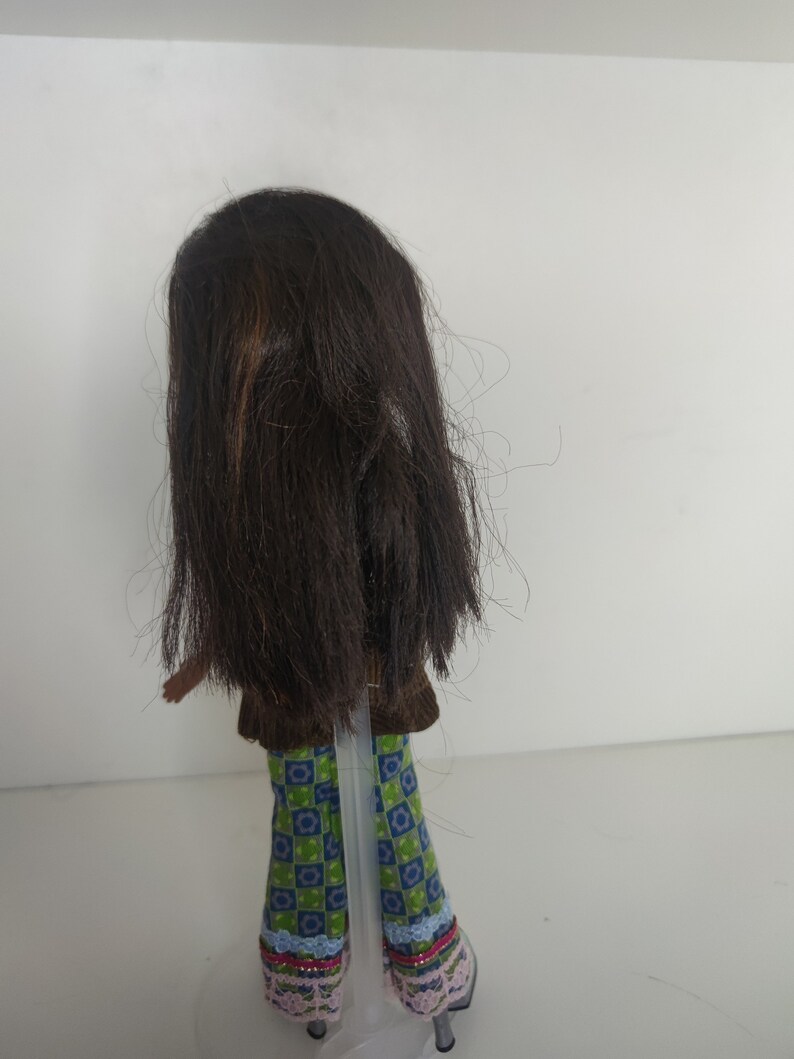 May include: A brown-haired doll wearing a brown vest, green and blue checkered pants with pink and blue trim, and pink shoes.