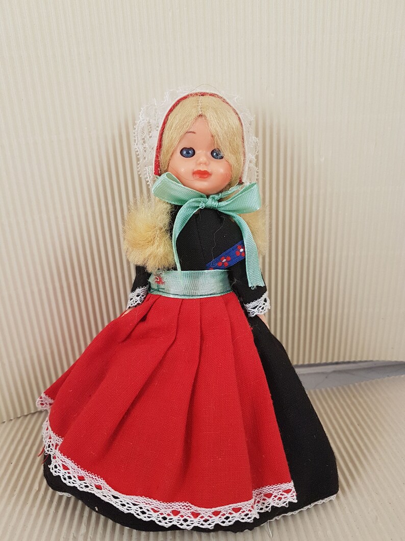 Puede incluir: Una peque&ntilde;a mu&ntilde;eca vestida con un traje tradicional holand&eacute;s. La mu&ntilde;eca tiene el pelo rubio, los ojos azules y un vestido rojo y negro con un borde de encaje blanco. La mu&ntilde;eca lleva una cofia de encaje blanco y una cinta verde alrededor del cuello.