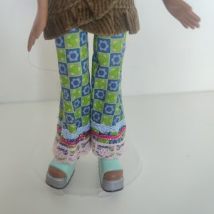 May include: A doll wearing a brown corduroy top with a blue and green patterned pants and light blue platform shoes.