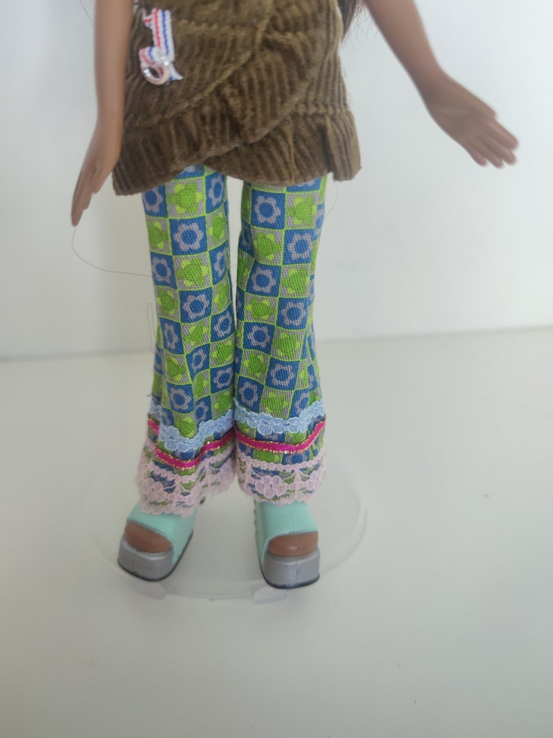 May include: A doll wearing a brown corduroy top with a blue and green patterned pants and light blue platform shoes.