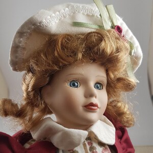 Puede incluir: Una mu&ntilde;eca de porcelana con el pelo rojo rizado que lleva un sombrero blanco con una cinta verde y un vestido rojo y blanco.