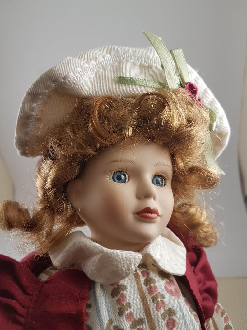 Puede incluir: Una mu&ntilde;eca de porcelana con el pelo rojo rizado que lleva un sombrero blanco con una cinta verde y un vestido rojo y blanco.