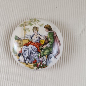 May include: A round ceramic trinket box with a painted scene of a couple in a garden setting. The woman is wearing a blue and white dress and the man is wearing a green and purple outfit. There is a small dog in the scene.
