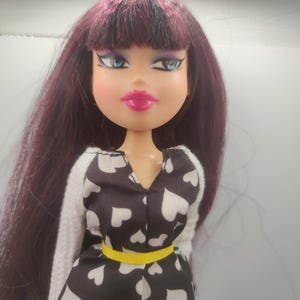 Pode incluir: Boneca Bratz com cabelo longo, roxo escuro e franja. A boneca tem olhos azuis, l&aacute;bios rosa e usa um macac&atilde;o preto e branco com estampa de cora&ccedil;&atilde;o e cinto amarelo. O traje da boneca inclui um su&eacute;ter branco e shorts cinza.