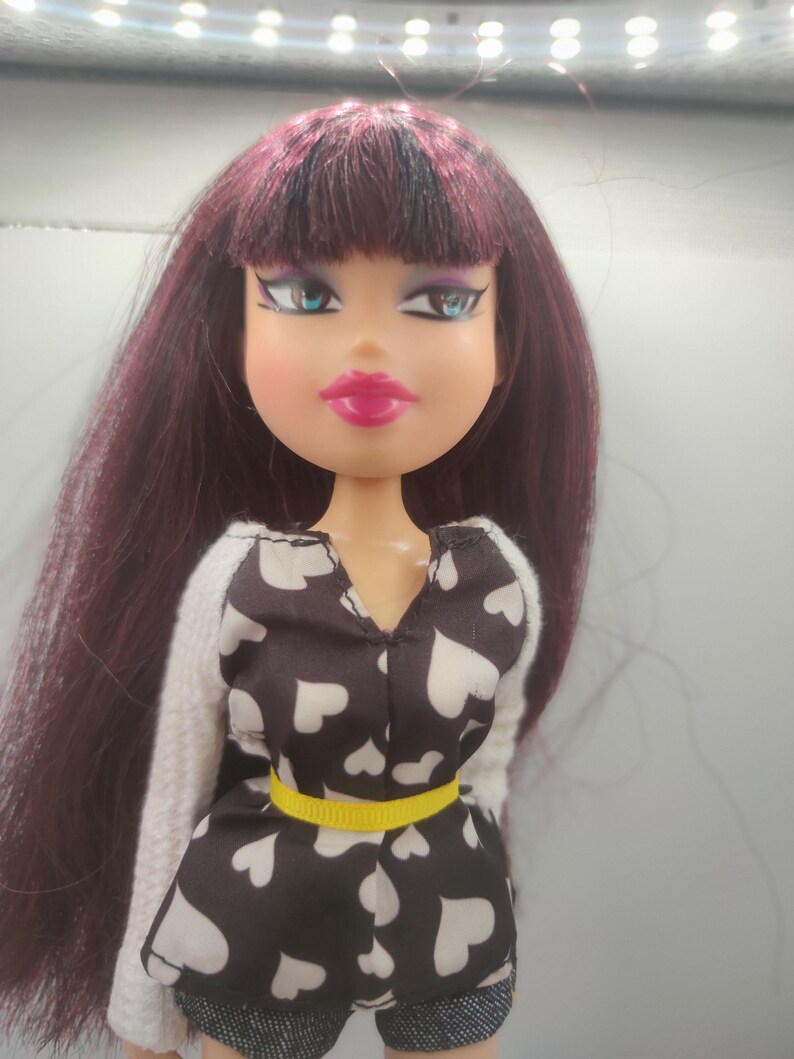 Pode incluir: Boneca Bratz com cabelo longo, roxo escuro e franja. A boneca tem olhos azuis, l&aacute;bios rosa e usa um macac&atilde;o preto e branco com estampa de cora&ccedil;&atilde;o e cinto amarelo. O traje da boneca inclui um su&eacute;ter branco e shorts cinza.