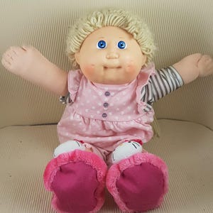 Peut inclure: Une poupée vintage avec des cheveux blonds en fil, des yeux bleus et des joues roses. La poupée porte une robe rose avec des motifs de cœurs blancs et un col à volants. La poupée a les bras tendus et des chaussures roses et floues.