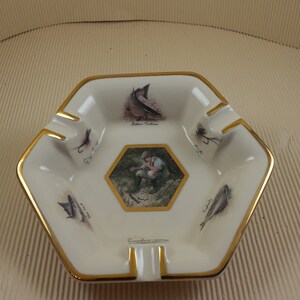 Peut inclure: Cendrier en céramique blanche avec un bord doré. Le cendrier présente une image centrale d'un pêcheur avec une canne à pêche et un poisson, entouré d'images de poissons et de mouches. Le texte "Fisherman's Collection" est imprimé sur le cendrier.