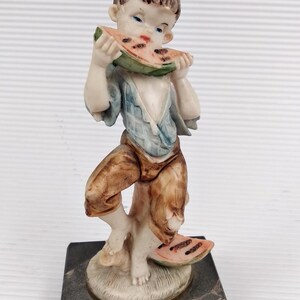 Puede incluir: Figura de cerámica de un joven comiendo sandía. La figura viste una camisa azul y pantalones marrones, de pie sobre una base decorativa. La rodaja de sandía es verde, roja y negra. La base es un cuadrado oscuro con aspecto de mármol.