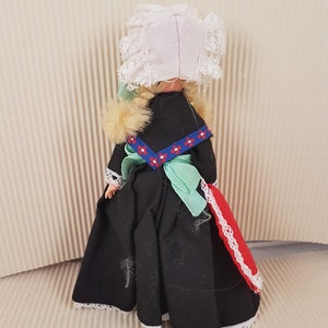 Puede incluir: Una peque&ntilde;a mu&ntilde;eca vestida con un vestido negro con ribete de encaje blanco, una faja verde y un gorro de encaje blanco. La mu&ntilde;eca tiene el pelo rubio y lleva una tela estampada roja y blanca en la parte posterior del vestido.