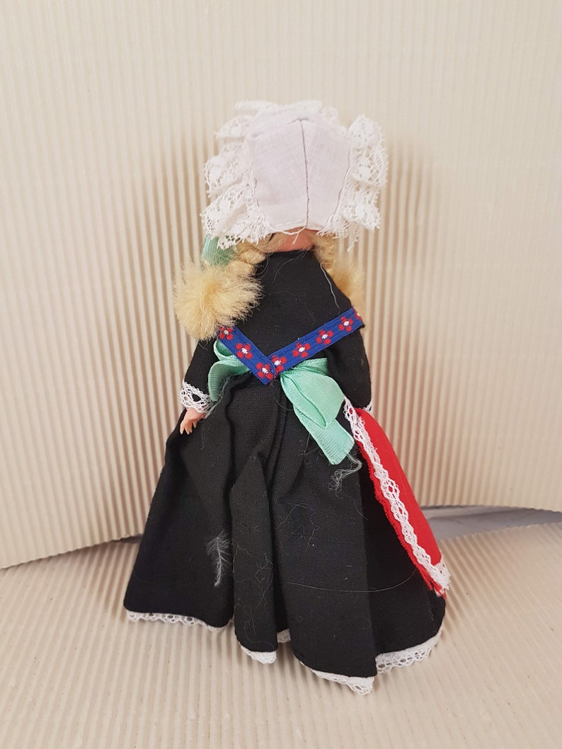 Puede incluir: Una peque&ntilde;a mu&ntilde;eca vestida con un vestido negro con ribete de encaje blanco, una faja verde y un gorro de encaje blanco. La mu&ntilde;eca tiene el pelo rubio y lleva una tela estampada roja y blanca en la parte posterior del vestido.