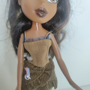 May include: A brown-skinned doll with long black hair wearing a brown corduroy skirt, a brown and gold corset, and green and blue checkered pants.